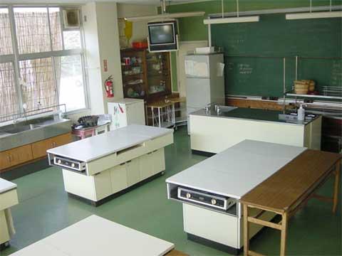 前方には黒板と冷蔵庫があり、白い天板の調理台が間隔をあけて設置されている調理室の写真