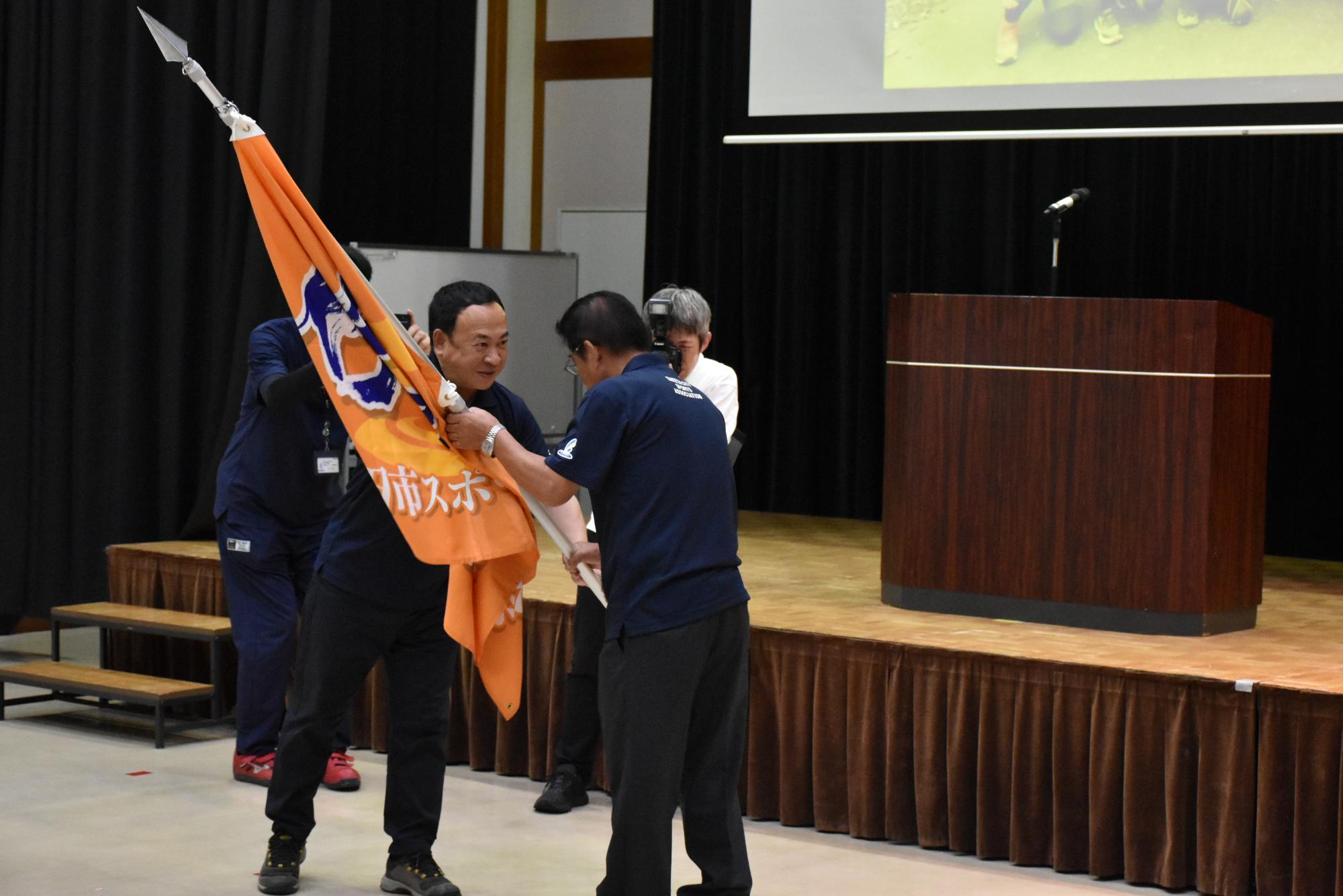 団旗推戴：土居 昌弘団長から本田 忠総監督（旗手）へ