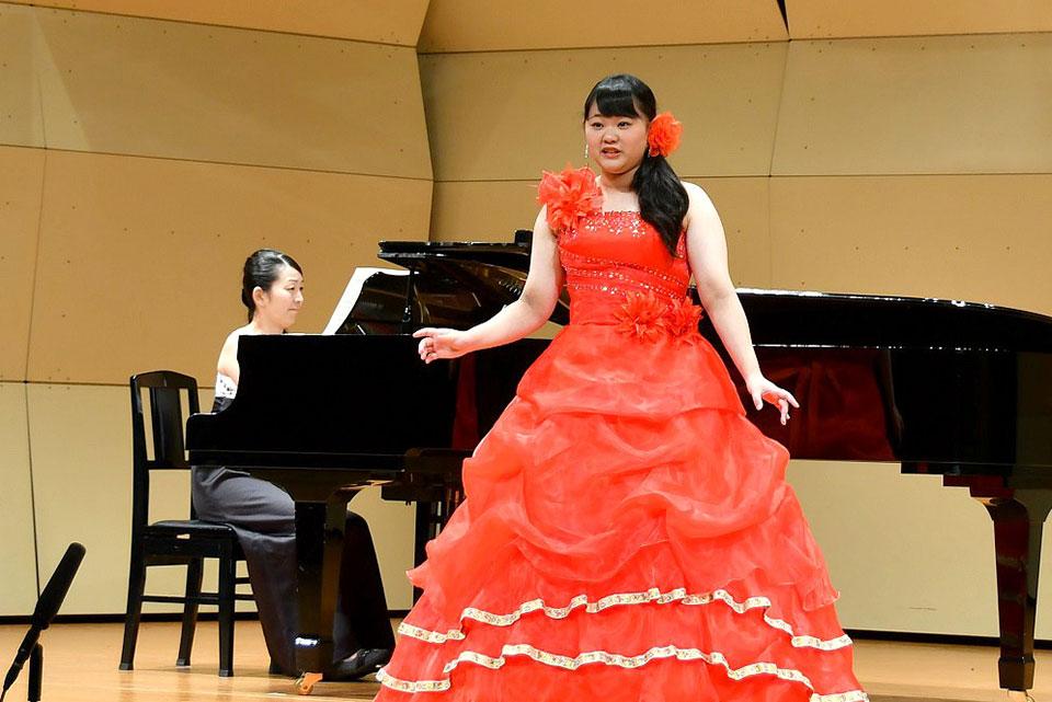 赤いドレスを着た塩谷梨奈さんがピアノの前で歌っている写真