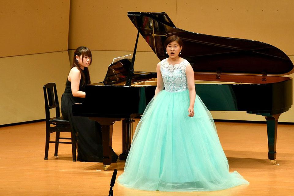 水色のドレスを着た中井香江さんがピアノの前で歌っている写真