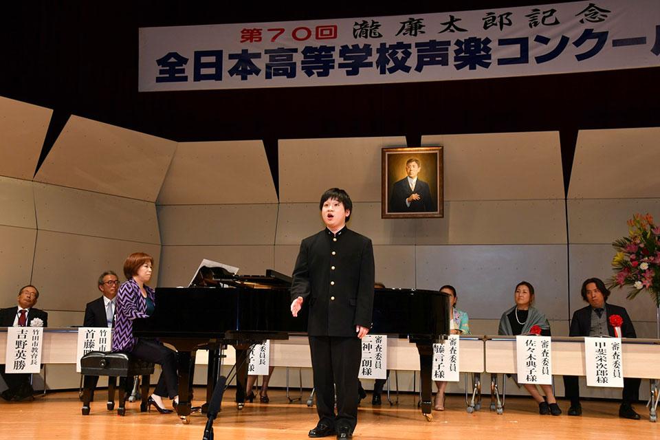 学生服を着た西谷亮良さんが審査委員の前でピアノに合わせて歌っている写真