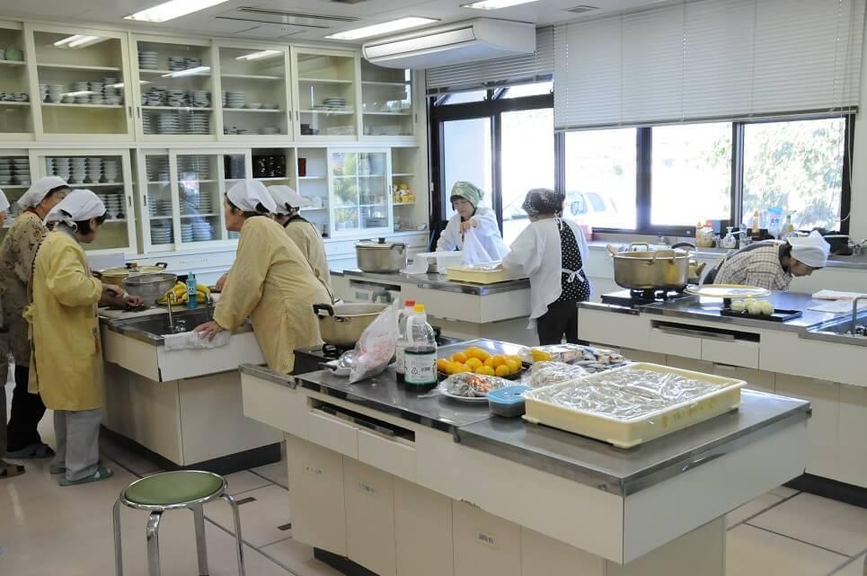 調理室の台に置かれた食材や大きな鍋と調理作業をしている、三角巾にエプロン姿の竹田市地域婦人団体連合会のメンバーの写真