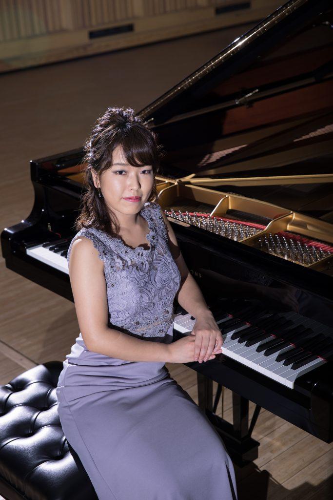 グランドピアノに左手をかけ、薄紫のドレスを着た古賀美代子さんの写真