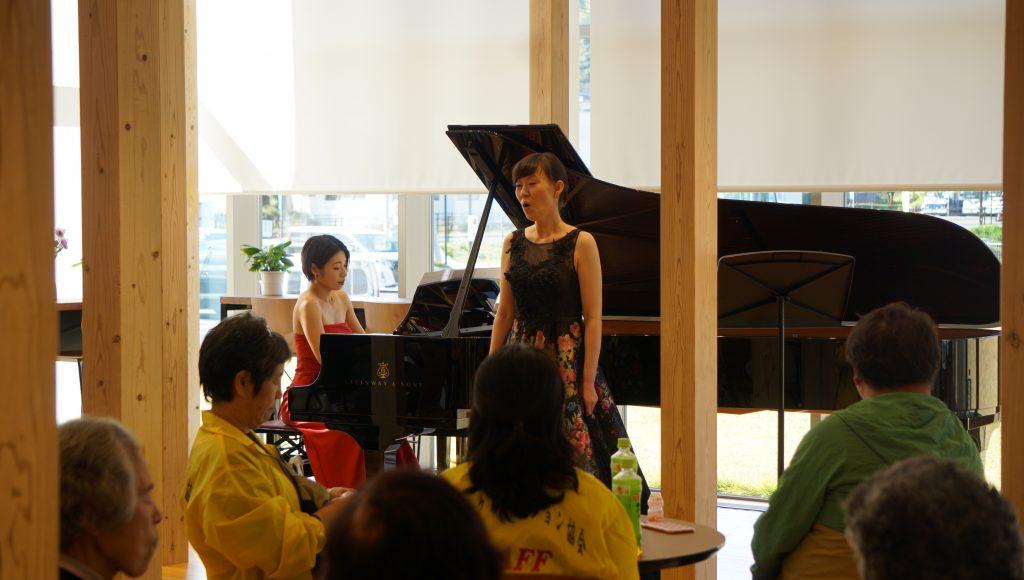 白いブラインドのあるラウンジで、歌う田島さんとピアノを演奏する小町さんと聞き入る観客たちの写真