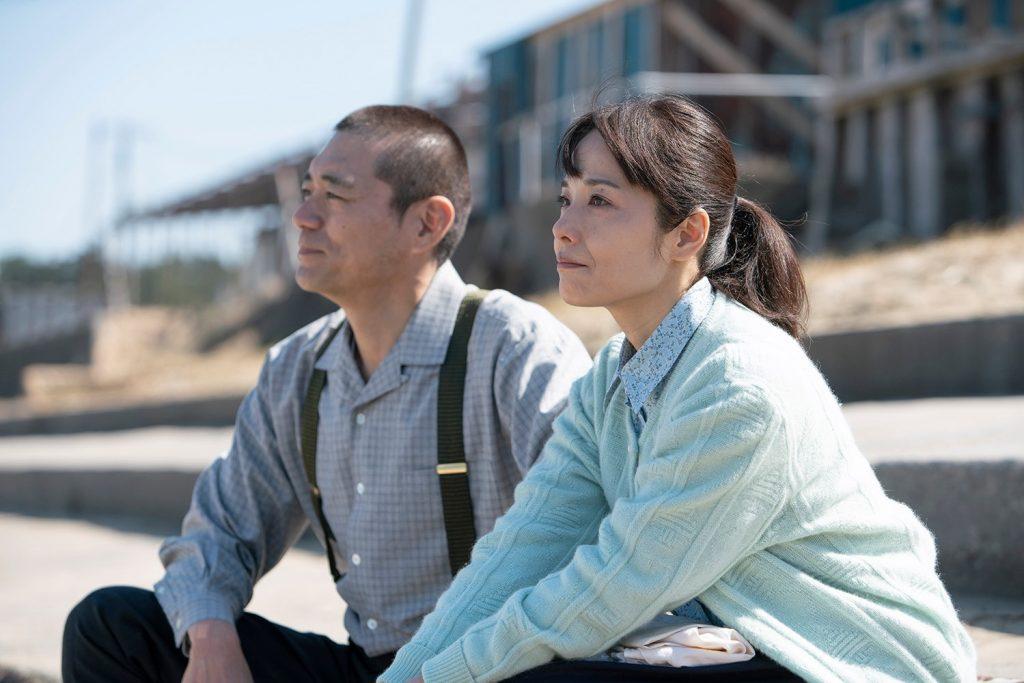 洋服姿の博多華丸と富田靖子が河川敷で座っている映画の一場面の写真