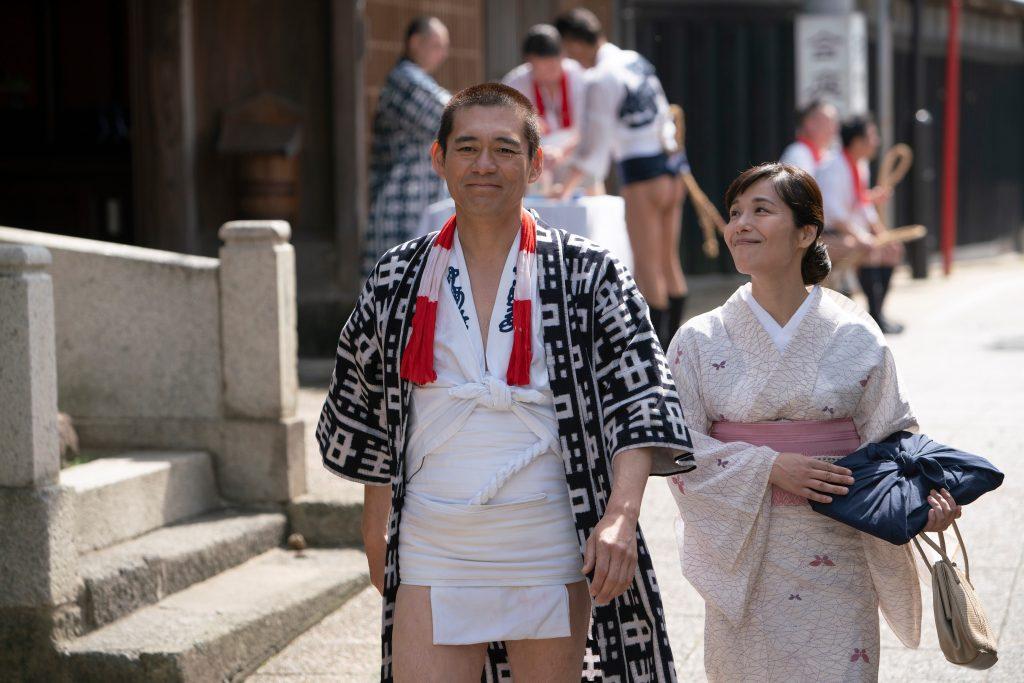 ふんどし姿に着物を羽織った博多華丸と風呂敷包みを抱えたピンクの着物を着た富田靖子の映画の一場面の写真